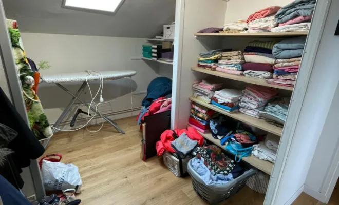 Description de l'Espace Dressing Avant Projet :  Découvrez l'espace dressing actuel de notre client, situé sous un élégant puits de lumière qui inonde la pièce de lumière naturelle. À gauche, un coin est dédié au repassage, ajoutant une touche fonctionnelle à cet espace. À droite, des étagères ouvertes bien remplies accueillent soigneusement une variété de vêtements et textiles, illustrant le besoin d'organisation et de rangement optimisés. Le sol en parquet clair ajoute une chaleur naturelle à l'ambiance. 